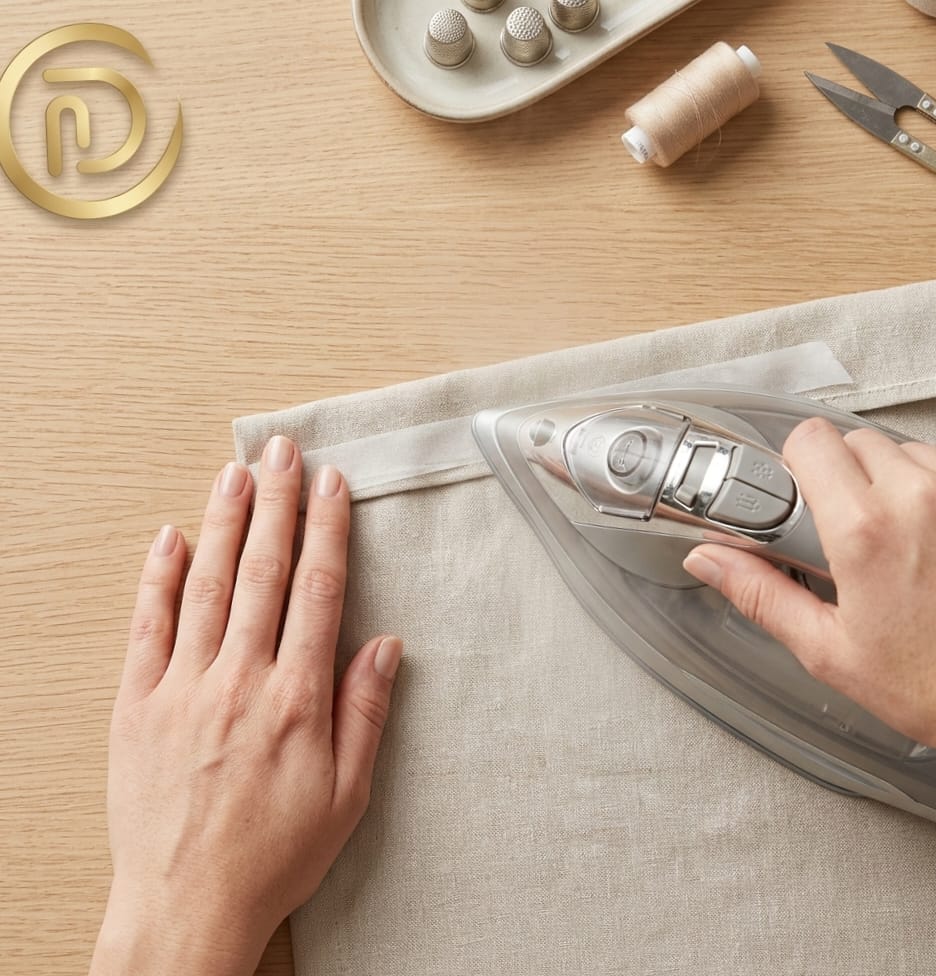 Close-up of hands using an iron to apply hem tape to cotton fabric for an invisible no-sew curtain hem.